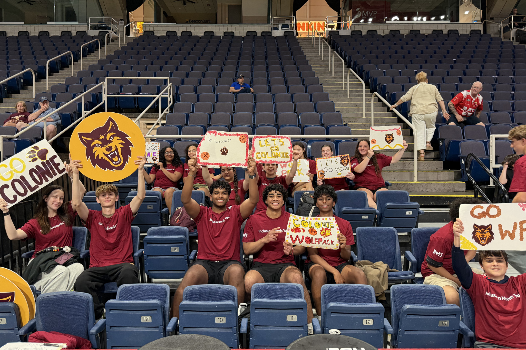 A group of students sit in arena seats wearing red shirts and holding up Go Colonie and Wolf Pack signs